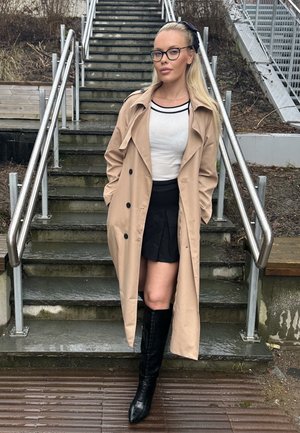 Trench beige con botones negros, llevado sobre una blusa blanca y una falda negra, combinado con botas negras de tacón hasta la rodilla, en un fondo de escalones de piedra.