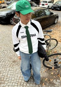 Chemise blanche à manches longues avec rayures noires, cravate verte et casquette verte. Jean bleu large et veste noire matelassée. Surface pavée avec des feuilles mortes.