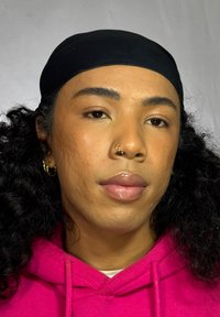 Personne aux cheveux noirs bouclés portant un foulard noir et un sweat à capuche rose, arborant un anneau de septum et des boucles d'oreilles en créole, avec une peau lisse.