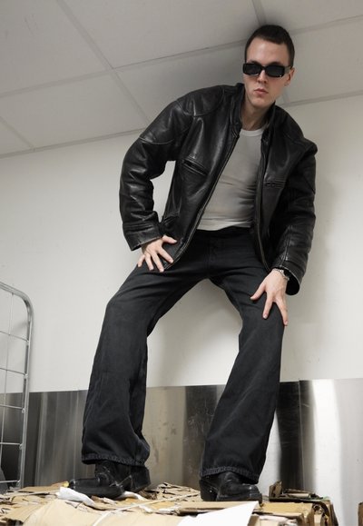 Chaqueta de cuero negra, camiseta gris y pantalones de campana negros llevados por una persona que está de pie sobre cajas de cartón; la persona lleva gafas de sol oscuras.