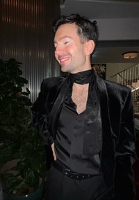 Blazer en velours noir avec col châle, associé à une chemise noire boutonnée comportant une cravate. Cadre intérieur avec de la verdure en arrière-plan.