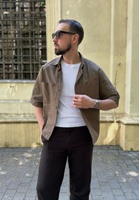 Bruin kortmouwen button-down shirt over een wit T-shirt, gecombineerd met zwarte broek. Accessoires zijn onder andere een zonnebril en een horloge.