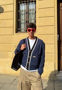 Granatowy kardigan z białymi akcentami, drewniane guziki, noszony na białej koszulce. Beżowe krótkie spodenki typu cargo i czarna torba na ramię, czerwone okulary przeciwsłoneczne.