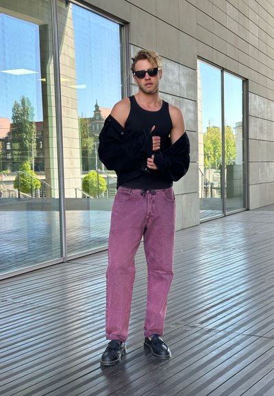 Chaqueta de mezclilla negra, top negro de tirantes y jeans rosas holgados. Zapatos negros. De pie sobre un suelo de madera con grandes ventanales de vidrio al fondo.