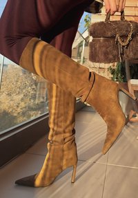 Stivali al ginocchio in camoscio marrone con punta a punta e tacco a spillo, caratterizzati da un design rilassato e una texture morbida. Accessorio: borsa in pelliccia.