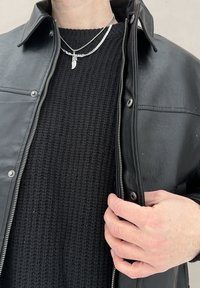 Homme boutonnant une veste en cuir noire par-dessus un pull en maille noire, portant deux colliers en argent dont un avec un pendentif en forme de plume.