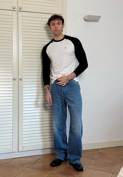 Hombre con pelo corto y rizado, que lleva una camisa blanca y negra de manga larga, vaqueros azules y zapatos negros, de pie en el interior junto a puertas blancas de armario.