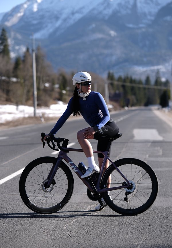 Cycliste portant un casque et des lunettes de soleil fait une pause sur la route avec son vélo, des montagnes enneigées et des pins en arrière-plan.