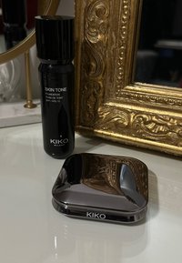 Sort rektangulær foundationflaske med pumpe mærket "Hudtone" SPF 15 ved siden af en blank mørk kompakt med "KIKO" logoet på en hvid overflade.