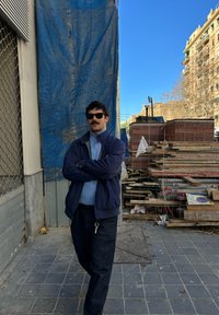 Egy ember karba tett kézzel áll egy kék ponyva előtt; napszemüveget, sötét dzsekit és kék inget visel. Körülötte láthatók építkezési anyagok.
