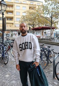 Szürke pulóver, melyen fekete "SOHO" felirat látható, sötétzöld nadrággal párosítva. A hátt�érben parkolt kerékpárok és őszi lombszerkezet látható.