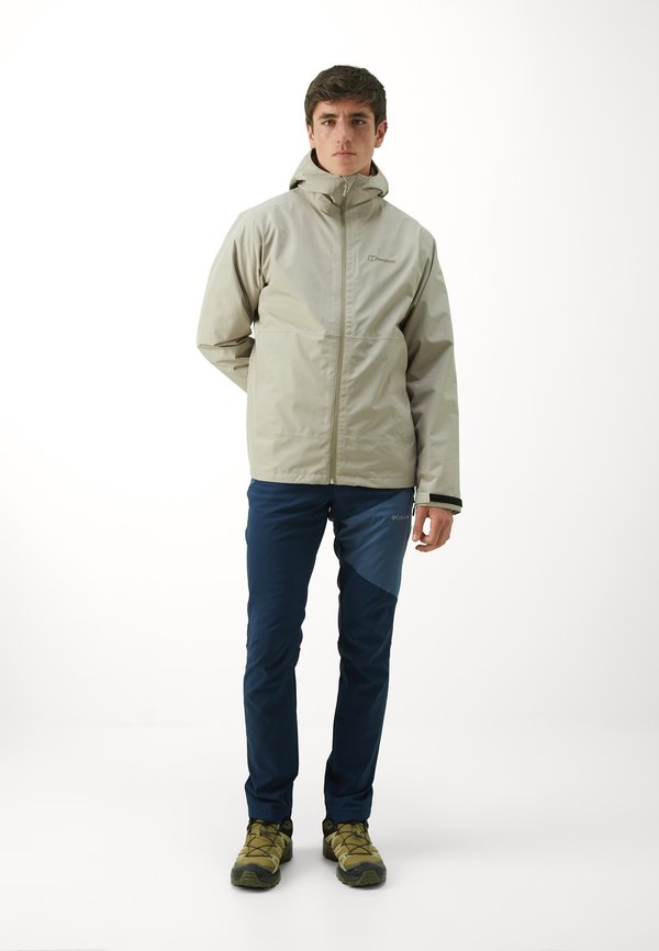 Giacca impermeabile colore beige chiaro con cappuccio, chiusura con zip e polsini elasticizzati. Abbinata a pantaloni blu scuro con design a pannelli e scarpe da trekking verdi.