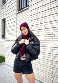 Schwarze, auffällige Jacke, kastanienbraune Mütze und Schal, beiges Oberteil und dunkle Shorts. Das Model posiert mit verschränkten Armen vor einer hellen Backsteinmauer.