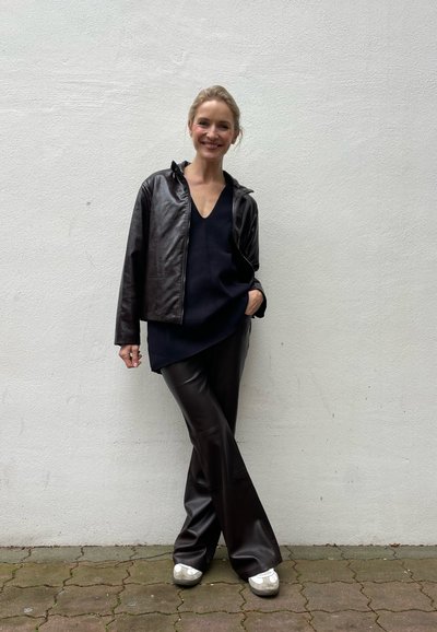 Chaqueta de cuero marrón, blusa de cuello en v oscura y pantalones de cuero sintético marrón, combinados con zapatos de color claro. De pie contra una pared blanca.