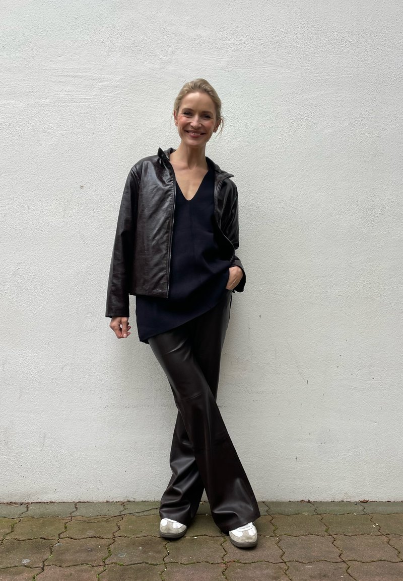 Pruun nahast jakk, tume V-kaelusega topp ja pruuni kunstnahast püksid, millele on lisatud heledat värvi jalanõusid. Seisab valge seina vastu.