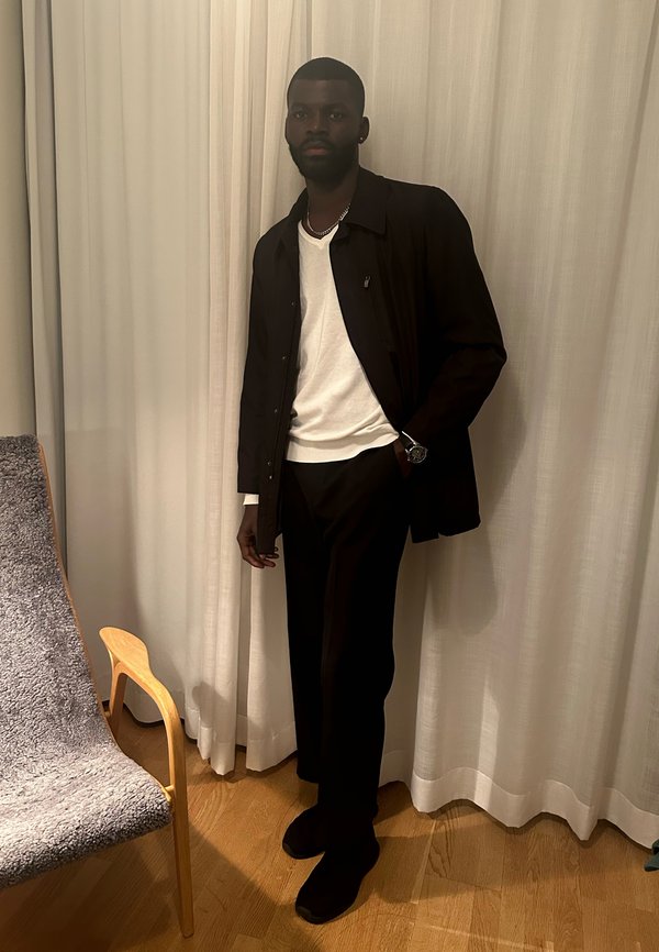 Mann trägt schwarze Jacke, weißes Hemd, schwarze Hose und Uhr, steht neben einem Holzstuhl mit grauem Stoff vor weißen Vorhängen.