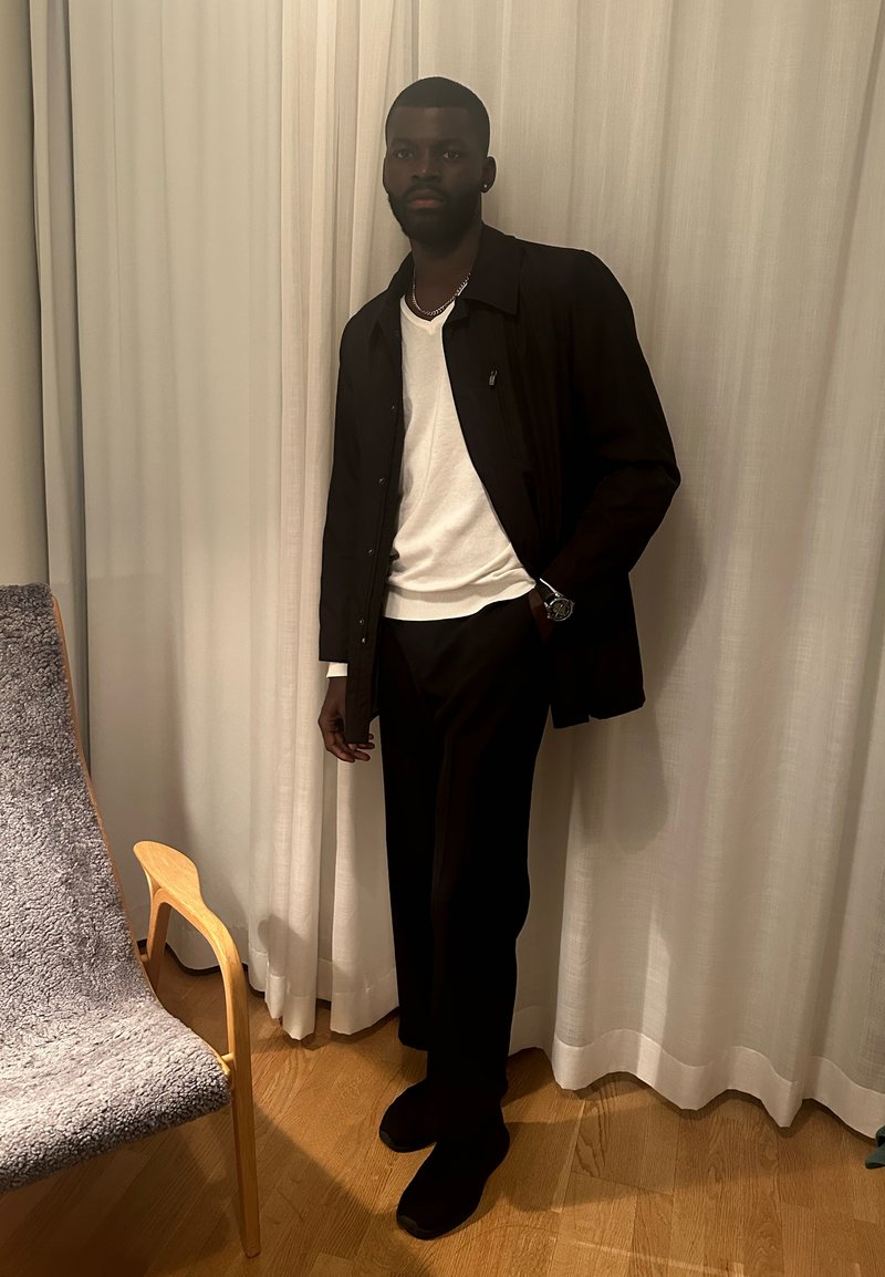 Άνδρας με μαύρο μπουφάν, λευκό πουκάμισο, μαύρο παντελόνι και ρολόι, στέκεται κοντά σε καρέκλα από ξύλο με γκρι ύφασμα μπροστά από λευκές κουρτίνες.