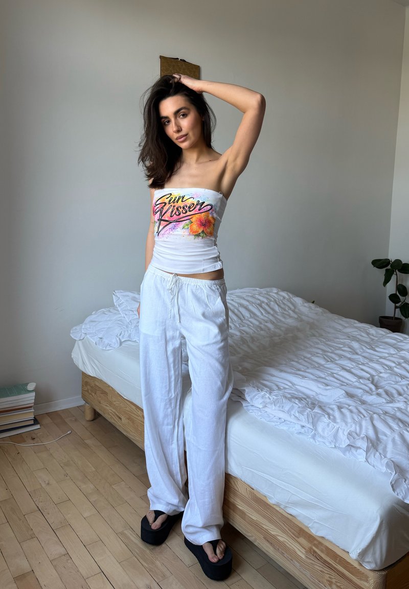 Femme aux cheveux foncés debout à côté d'un lit, portant un top tube blanc avec le texte «Sun Kisser», un pantalon blanc ample et des sandales à plateforme noires.