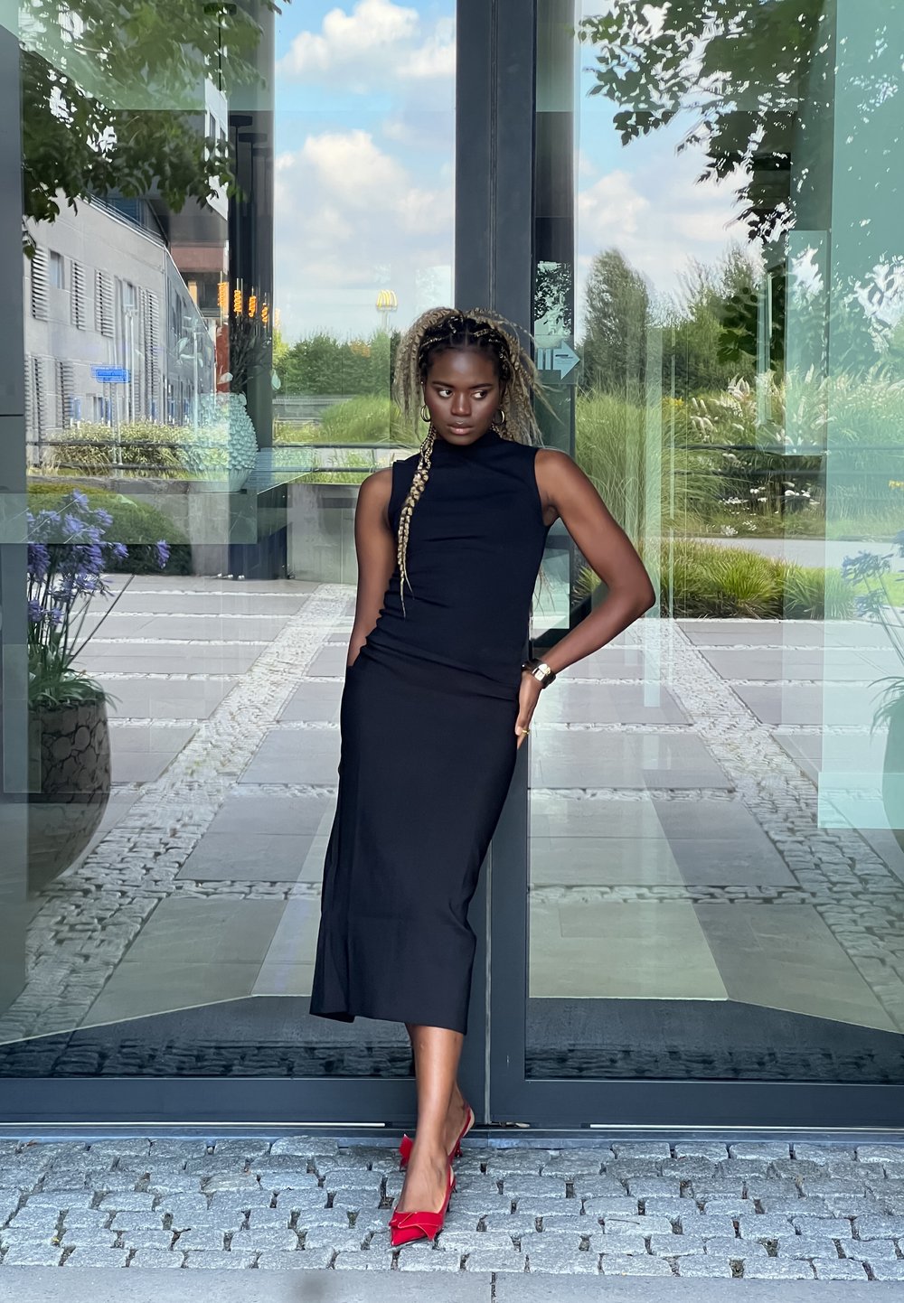 Μαύρο αμάνικο φόρεμα με ψηλό λαιμόκοψη και εφαρμοστή σιλουέτα, που διαθέτει πλευρικό άνοιγμα και συνδυάζεται με έντονα κόκκινα ψηλοτάκουνα.