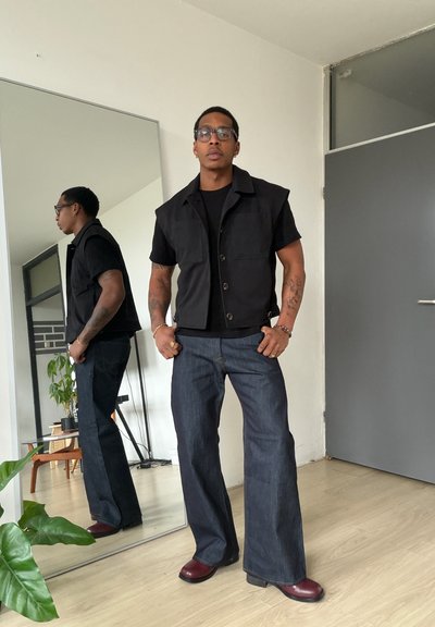 Hombre con gafas, chaqueta negra sin mangas, camisa negra, vaqueros de pierna ancha y zapatos marrones de pie frente a un espejo en una habitación minimalista.