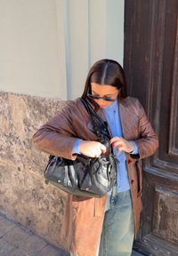 Femme portant des lunettes de soleil et un manteau en cuir marron, ouvrant un sac à main noir debout près d'un mur de pierre et d'une porte en bois.