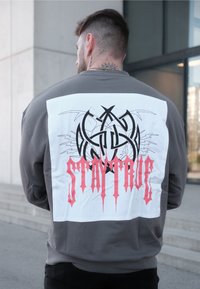 Sivá košeľa s dlhým rukávom a výrazným bielym zadným panelom, ktorý obsahuje čierny grafický dizajn a červený text "STAY TRUE".