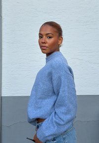 Jeune femme aux cheveux tirés en arrière, portant un pull bleu et un jean, debout de profil contre un mur texturé blanc et gris.