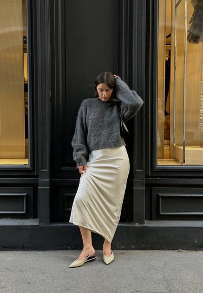 Suéter gris peludo y corto, falda midi de satén crema y zapatos de tacón bajo beige, con un bolso negro minimalista contra un fondo oscuro.