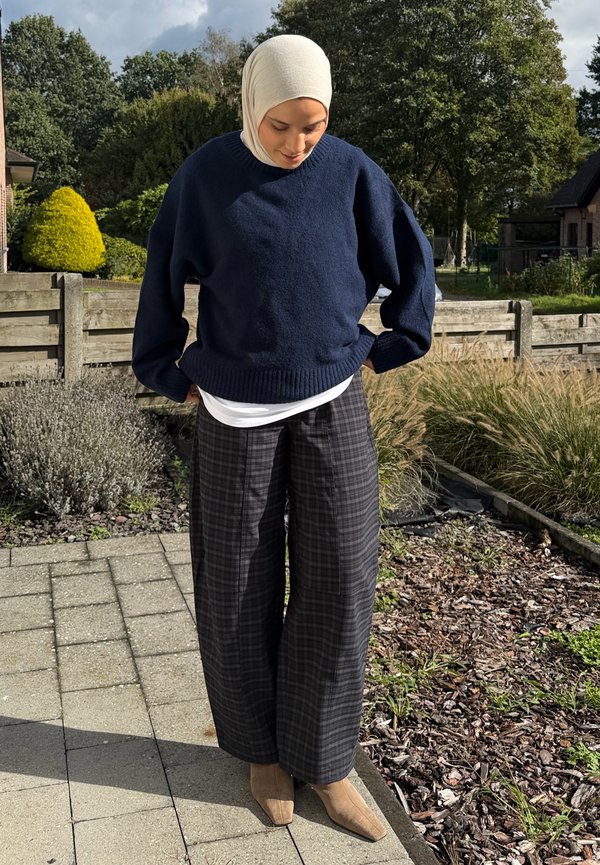 Une femme portant un hijab couleur crème, un pull bleu marine, un pantalon à carreaux et des bottines beige-brun se tient dehors, regarde vers le bas et ajuste son pull.