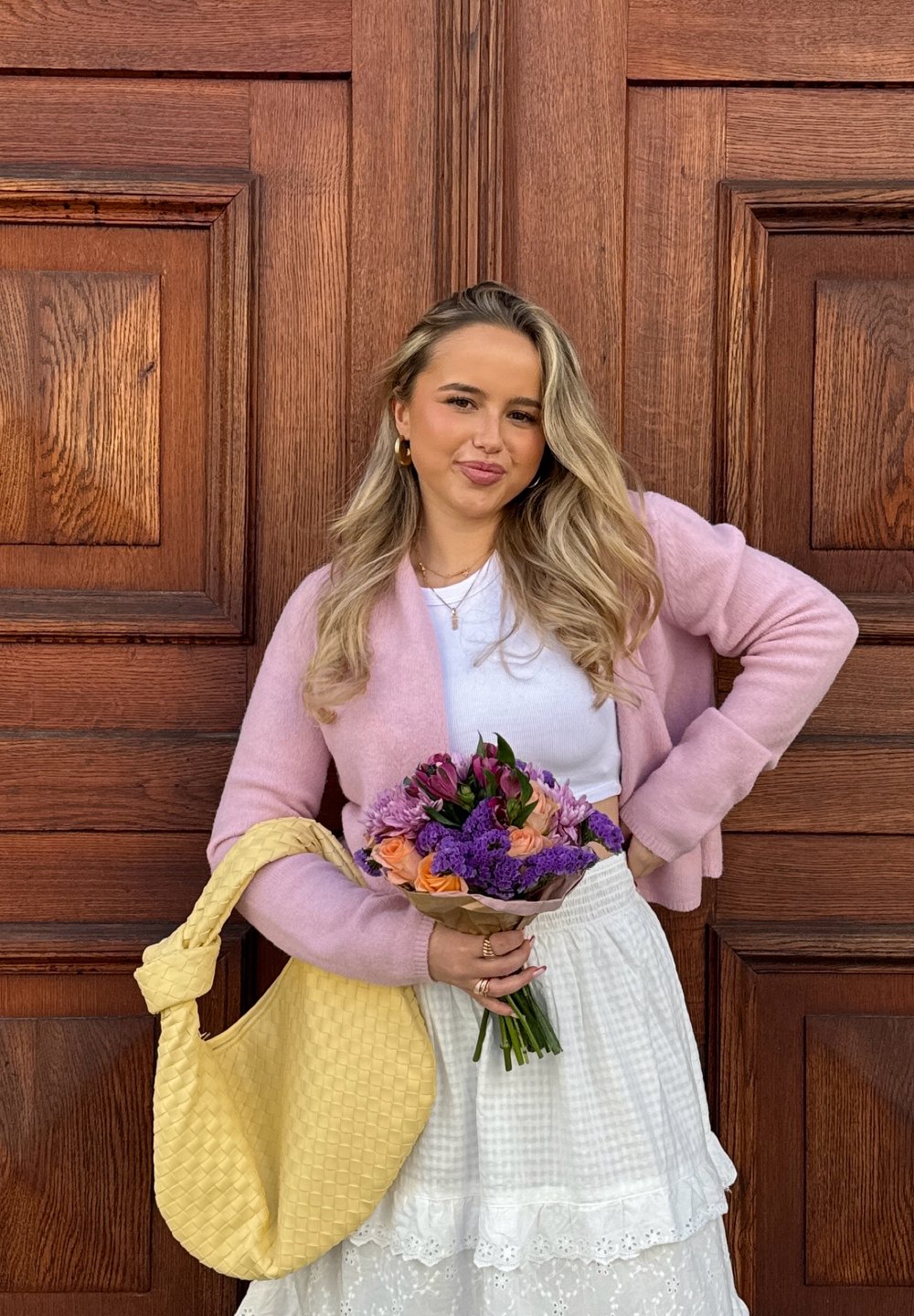 Helle rosa gestrickte Strickjacke über einem weißen Oberteil, kombiniert mit einem weißen, mehrlagigen Rock. Hält einen Blumenstrauß aus bunten Blumen und einer gelben geflochtenen Tasche.