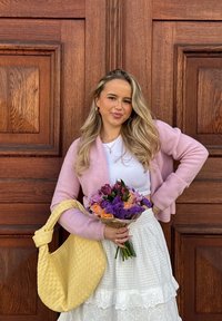 Helle rosa gestrickte Strickjacke über einem weißen Oberteil, kombiniert mit einem weißen, mehrlagigen Rock. Hält einen Blumenstrauß aus bunten Blumen und einer gelben geflochtenen Tasche.