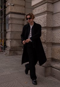 Melnā gara mētelis pāri baltai T-kreklam un svītrainai kreklai. Melnas bikses un spīdīgas kurpes. Valkā saulesbrilles, staigājot pie teksturētas akmens sienas.