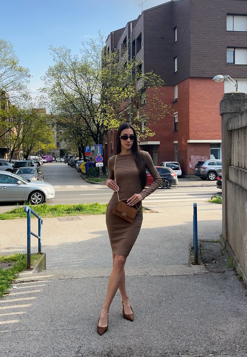 Femme en robe marron moulante et talons posant sur un trottoir urbain avec des arbres, des voitures stationnées et des bâtiments en briques en arrière-plan par une journée ensoleillée.