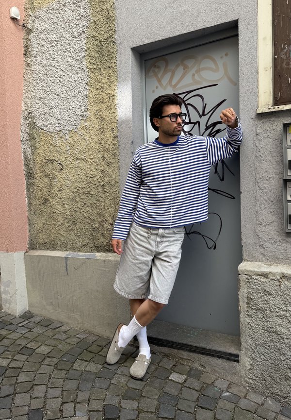 Bărbat purtând ochelari, cămașă cu mâneci lungi în dungi și pantaloni scurți, sprijinit de o ușă acoperită cu graffiti într-o stradă îngustă pietruită.
