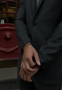 Szary garniturowy żakiet w prążki z guzikami, noszony na koszuli. Na nadgarstku widoczna zielona bransoletka z małymi akcentami. Czerwone tło.