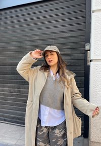 Cappotto oversized beige con tasche a toppa, maglione grigio lavorato a maglia, camicia bianca e pantaloni in camouflage. Cappellino camo grigio. Sfondo con una porta da garage nera.