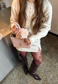 Femme en pull crème boutonné et jupe, tenant un sac à main rose avec une fleur, portant des collants en dentelle bordeaux et des bottes marron jusqu'aux genoux.