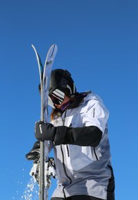 Des skieurs tenant des skis à double spatule, portant une veste grise et blanche, des gants noirs et un casque avec des lunettes à miroir, contre un ciel bleu clair.