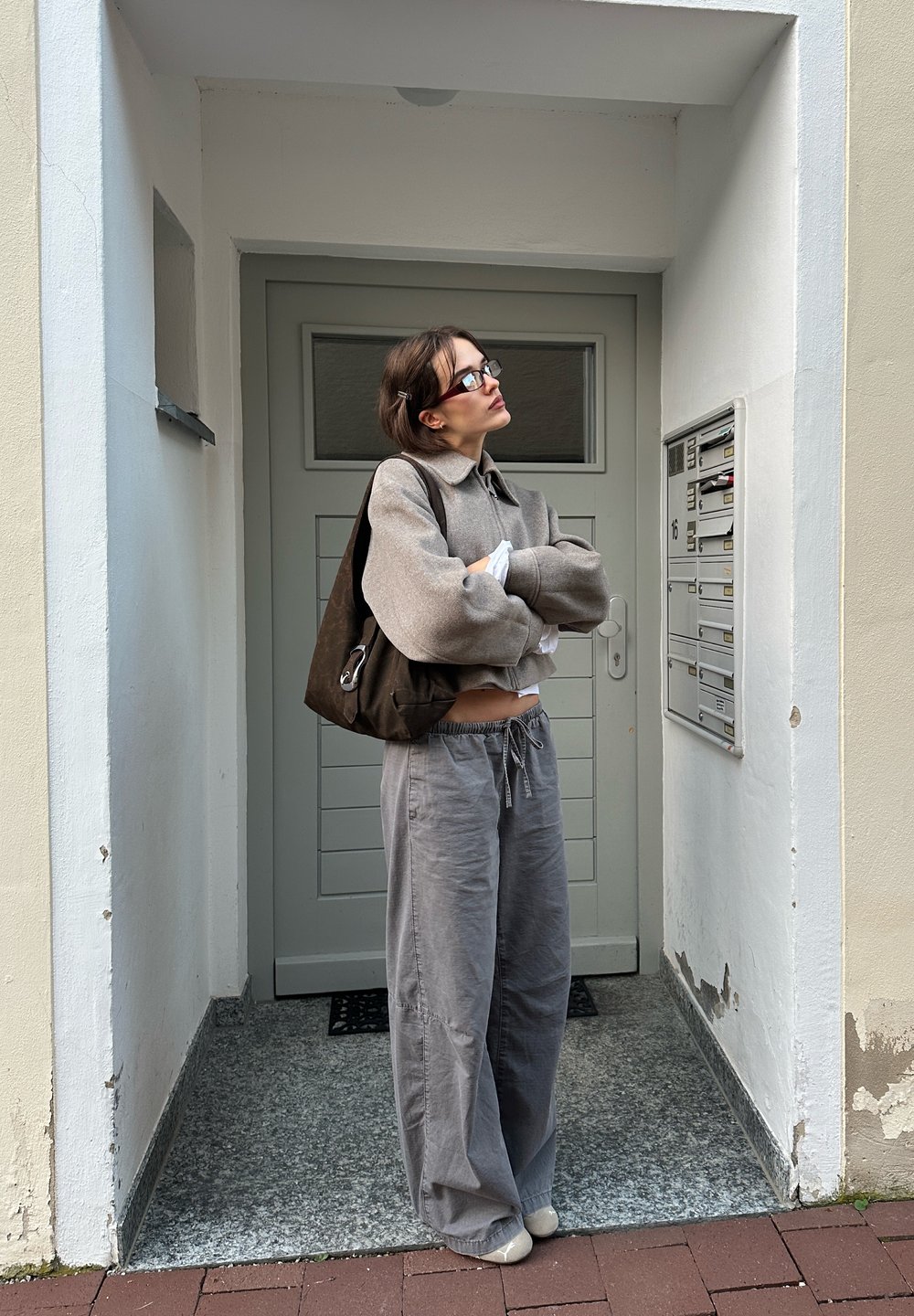 Szary, oversize sweter z kołnierzykiem, w połączeniu z luźnymi szarymi spodniami. Brązowa torba na ramię i minimalistyczne czarne okulary, na tle jasnej ściany.
