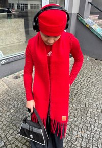 Cachecol de lã vermelho com franjas, blusa de manga longa vermelha a condizer, auscultadores pretos e uma pequena bolsa preta com detalhes e logotipo brancos, segurada na mão.