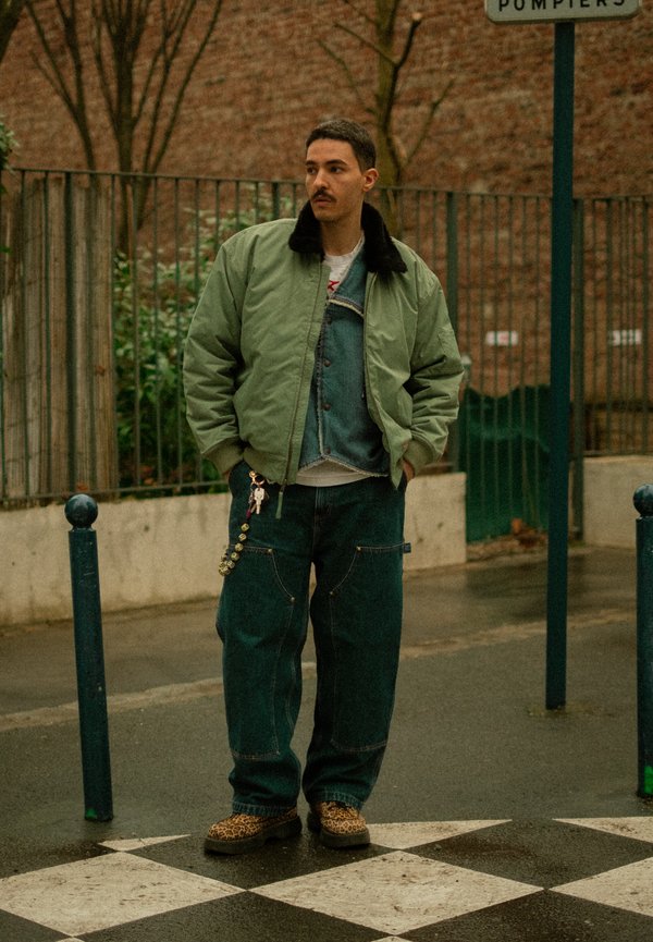 Man met snor die een groene bomberjas, een spijkervest, losse jeans en schoenen met luipaardprint draagt, staand op een geruite bestrating buiten.