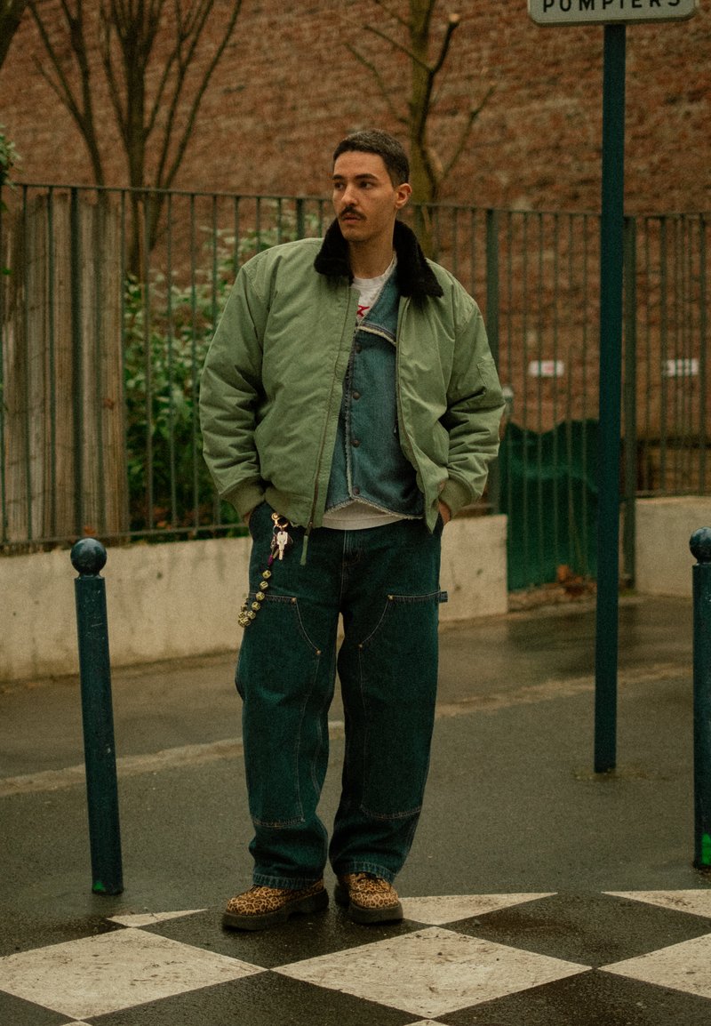 Homme avec une moustache portant une veste bomber verte, un gilet en jean, un jean large et des chaussures à motif léopard, debout sur un pavement à damier en plein air.