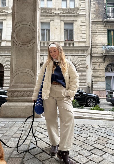 Joven mujer con chaqueta color crema y pantalones beige paseando a un perro por una calle de la ciudad con edificios históricos y coches al fondo.