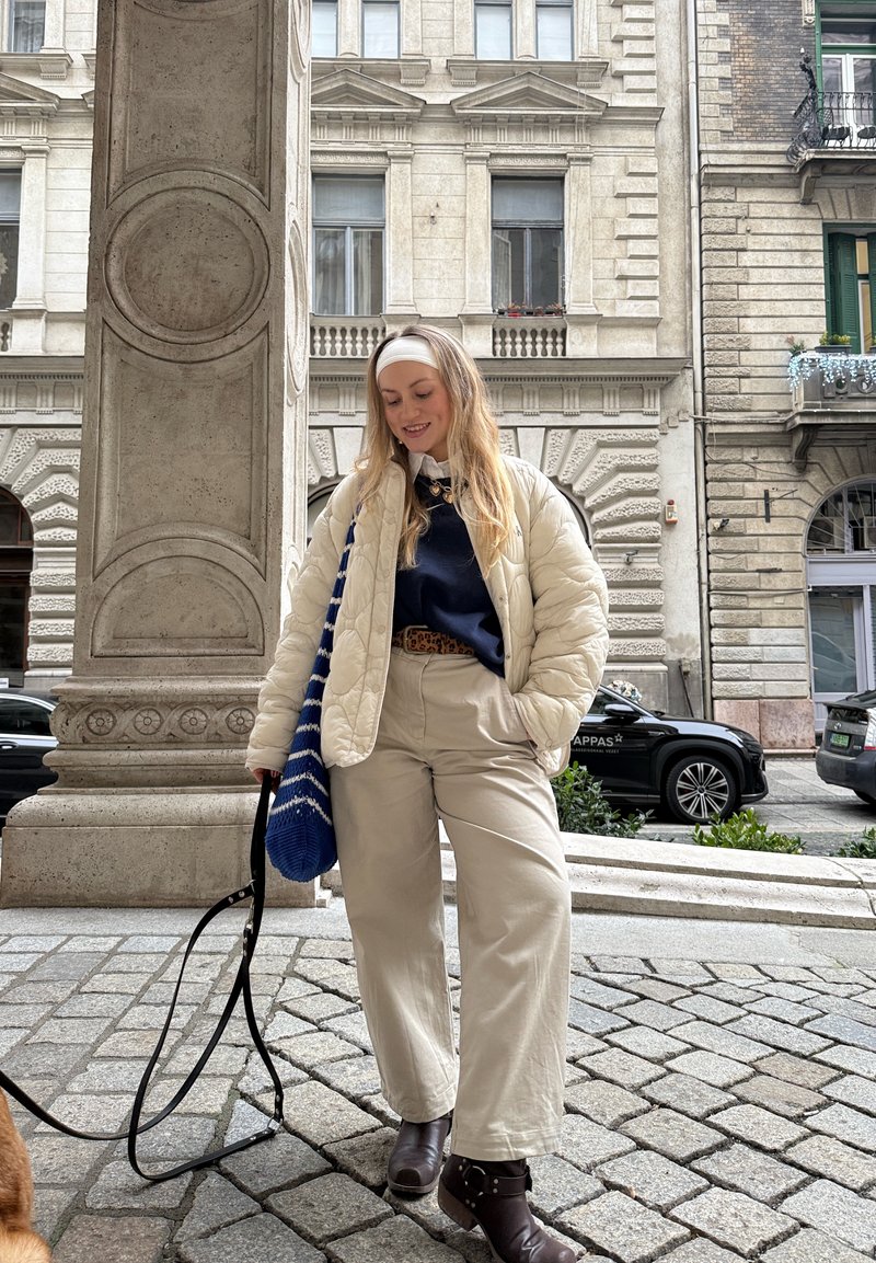 Jeune femme en veste crème et pantalon beige promenant un chien dans une rue de la ville avec des bâtiments historiques et des voitures en arrière-plan.
