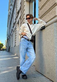 Ανοιχτό γκρι πουλόβερ, φαρδιά μπλε τζιν παντελόνια, μαύρα χοντρά παπούτσια, καφέ τσάντα ώμου, στέκεται μπροστά από ένα κτίριο με ανακλαστικό παράθυρο.