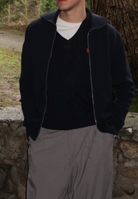 Veste zippée bleu marine portée sur un pull foncé, associée à un pantalon gris clair. La tenue présente un design épuré et décontracté avec une coupe ample.