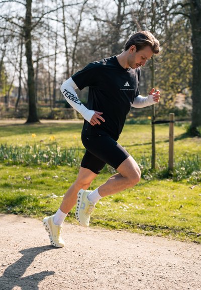 Hombre vestido con ropa deportiva negra y zapatillas blancas corriendo por un camino de grava en un parque con árboles y césped.