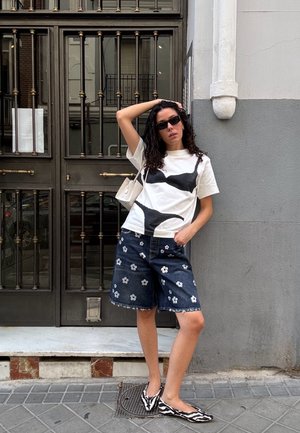 Mujer con cabello largo y rizado, usando gafas de sol negras, camiseta gráfica blanca, pantalones cortos de mezclilla con flores, zapatos planos de rayas de cebra y un pequeño bolso blanco.