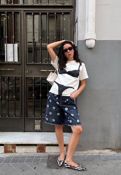 Mujer con cabello largo y rizado, usando gafas de sol negras, camiseta gráfica blanca, pantalones cortos de mezclilla con flores, zapatos planos de rayas de cebra y un pequeño bolso blanco.