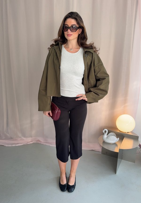 Une femme porte une veste vert olive, un haut blanc, un pantalon noir court, des lunettes de soleil et des chaussures plates. Elle tient une pochette bordeaux près d’une lampe de table allumée.